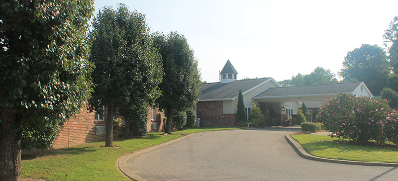 A single-story brick building with a white cupola and covered entrance, surrounded by trees and neatly maintained landscaping, with a driveway leading to the entrance.