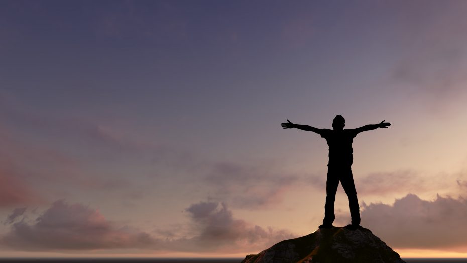 A person stands on a rock with arms outstretched against a colorful sunset sky.