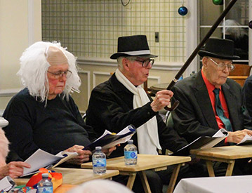 Three elderly individuals, two wearing black hats and one with a white wig, are seated in a row at a meeting, reading from their papers. A gavel and water bottles are present on the table in front of them.