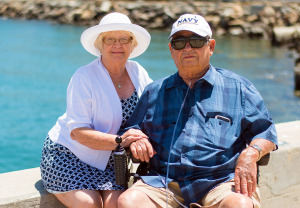 An elderly couple, with the woman wearing a white hat and the man in a navy cap, sit together on a bench by a waterfront on a sunny day.