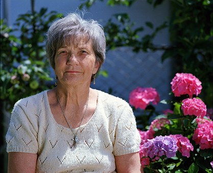 An elderly woman with short gray hair sits outdoors, wearing a white knitted top and a necklace. Pink and purple hydrangeas are in the background.