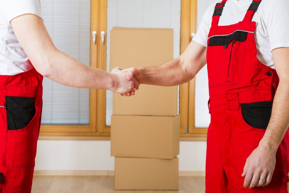 Two individuals in red work uniforms shake hands in front of a stack of cardboard boxes and large windows with blinds.