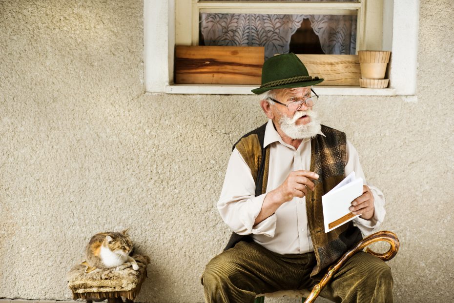 An elderly man wearing a green hat and traditional vest is seated outside a building, holding papers and a cane. A cat is curled up on a stool beside him.