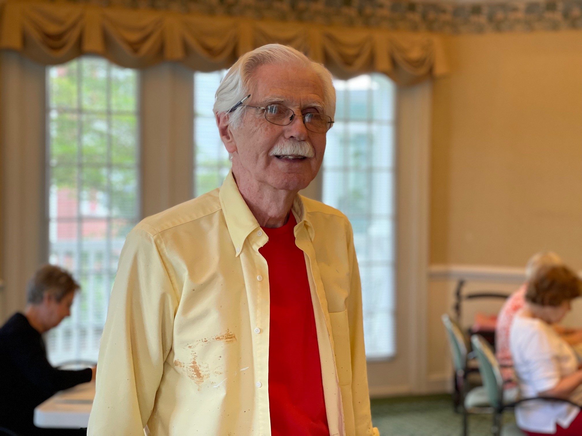 An elderly man with white hair and a mustache stands indoors wearing a yellow shirt over a red t-shirt. In the background, other people are seated, engaged in different activities, perhaps attending his painting classes.