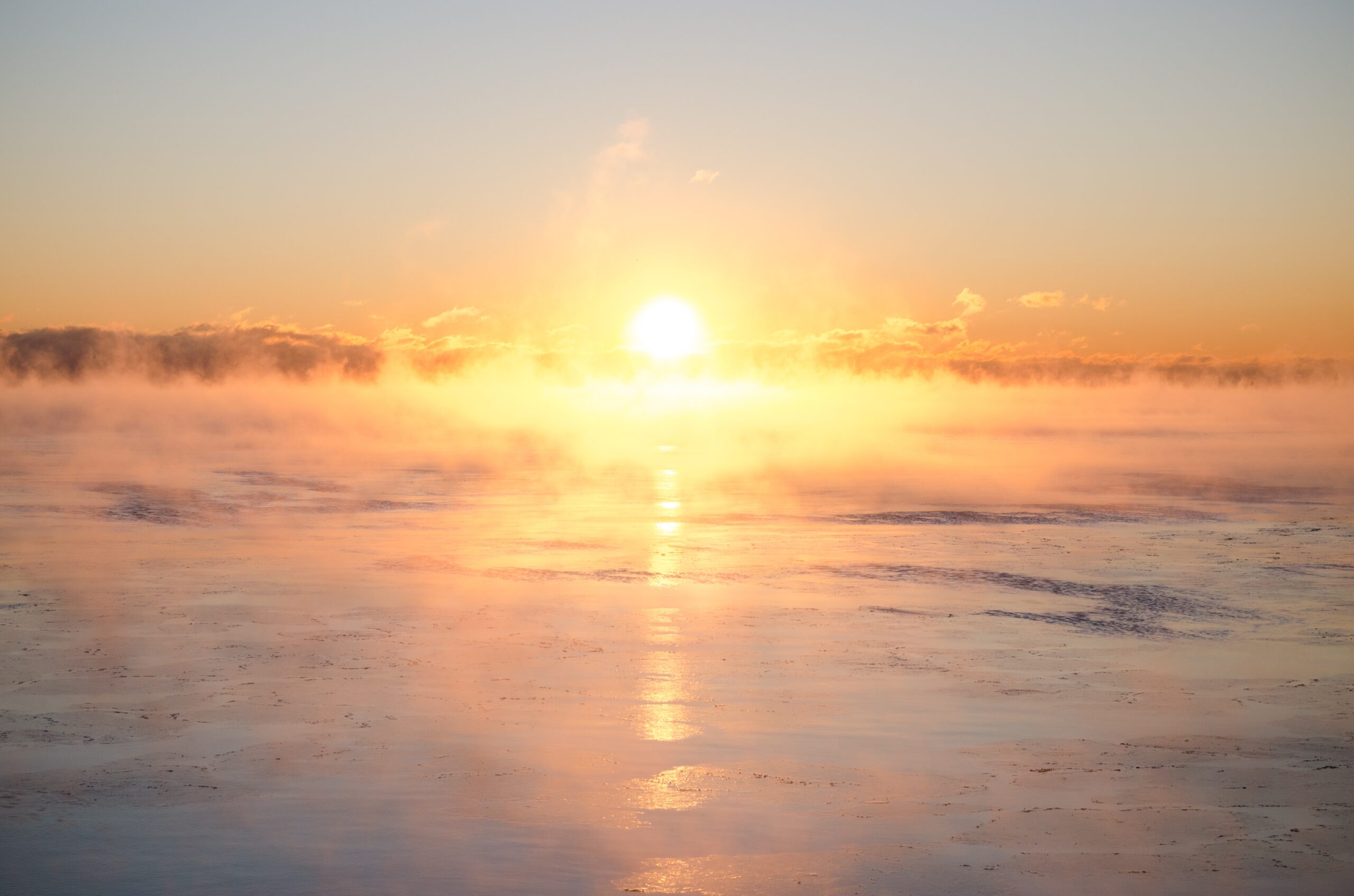 A sunrise over a calm body of water covered with mist, with warm orange and yellow hues reflecting on the water's surface.