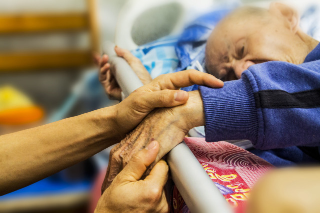 An elderly person lies in bed while another person gently holds their arm, offering comfort and care.