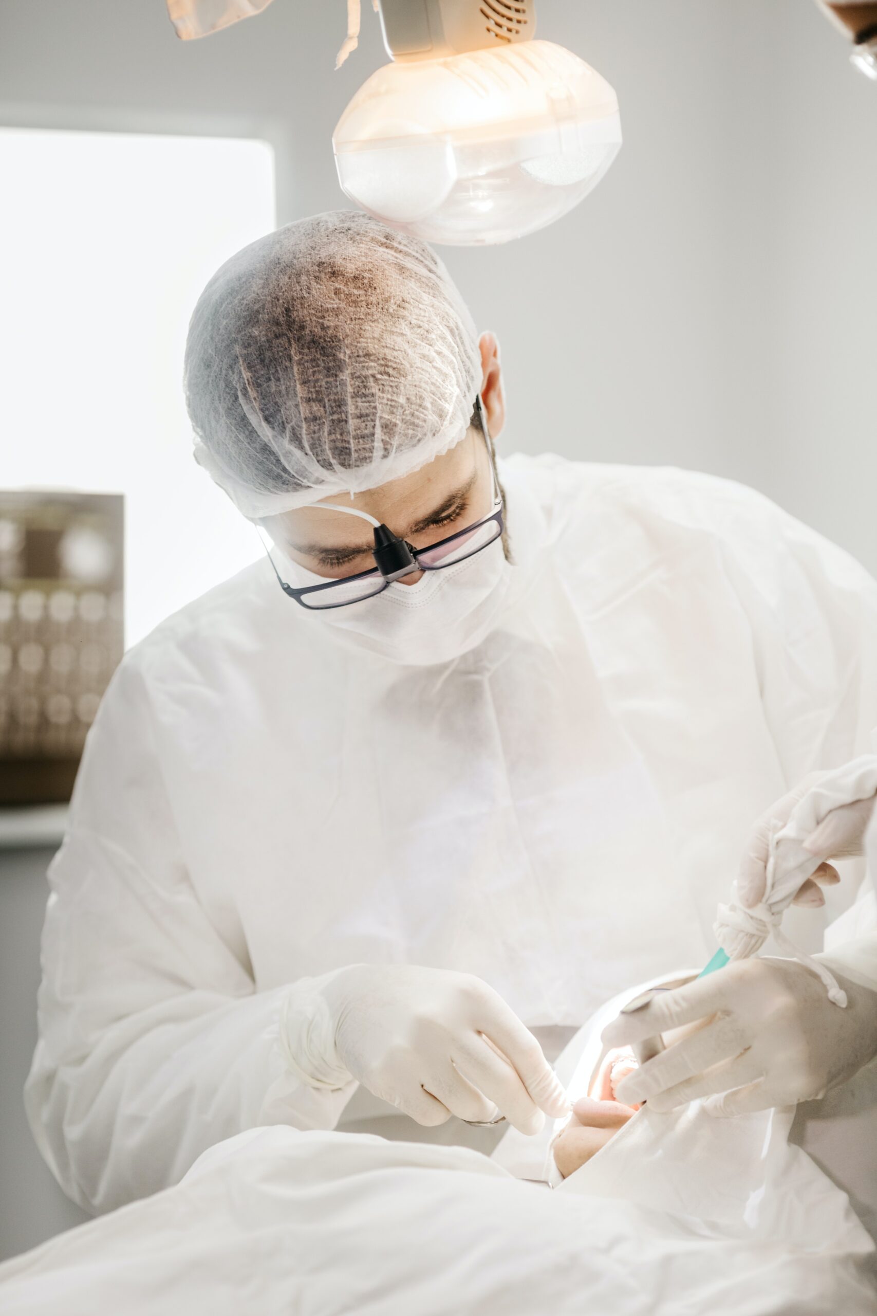 A dentist wearing a protective suit, mask, hairnet, and magnifying glasses performs a procedure on a patient in a clinical setting.