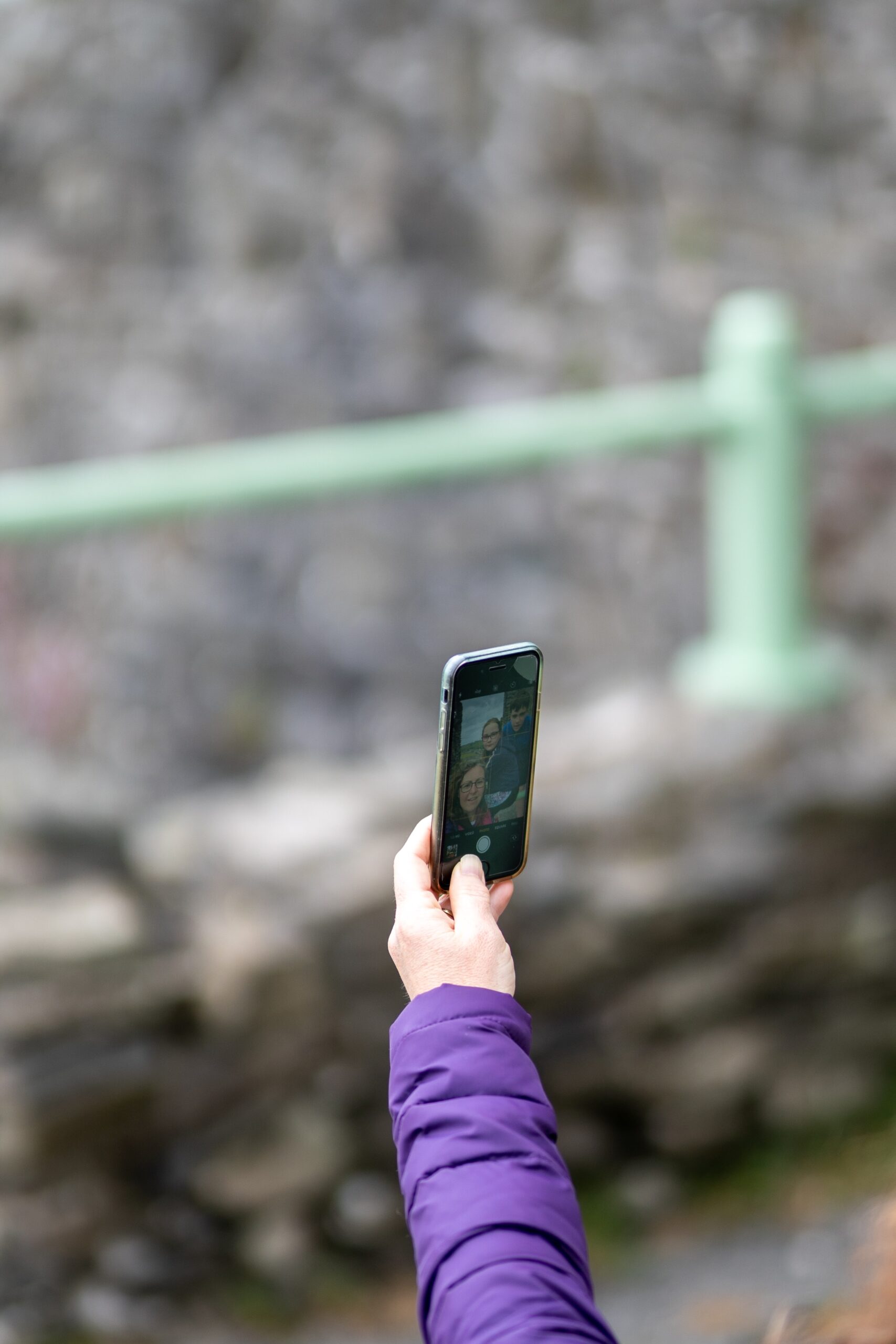 A person wearing a purple jacket holds up a smartphone, taking a selfie with three people visible on the phone screen, with a blurred background of a green railing and rocks.