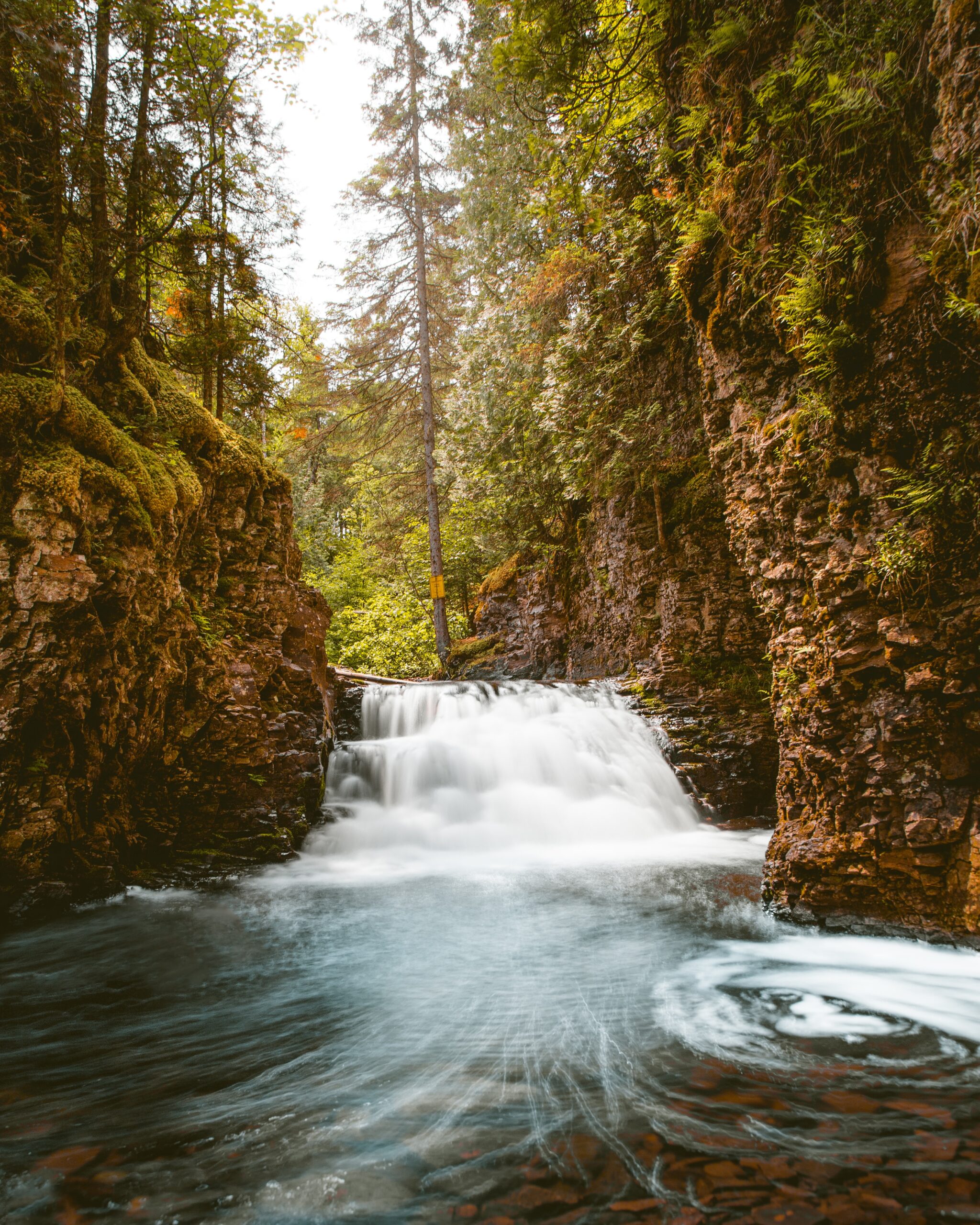 A small waterfall flows between two rocky cliffs with lush green foliage, into a swirling pool of water below in a forest setting.