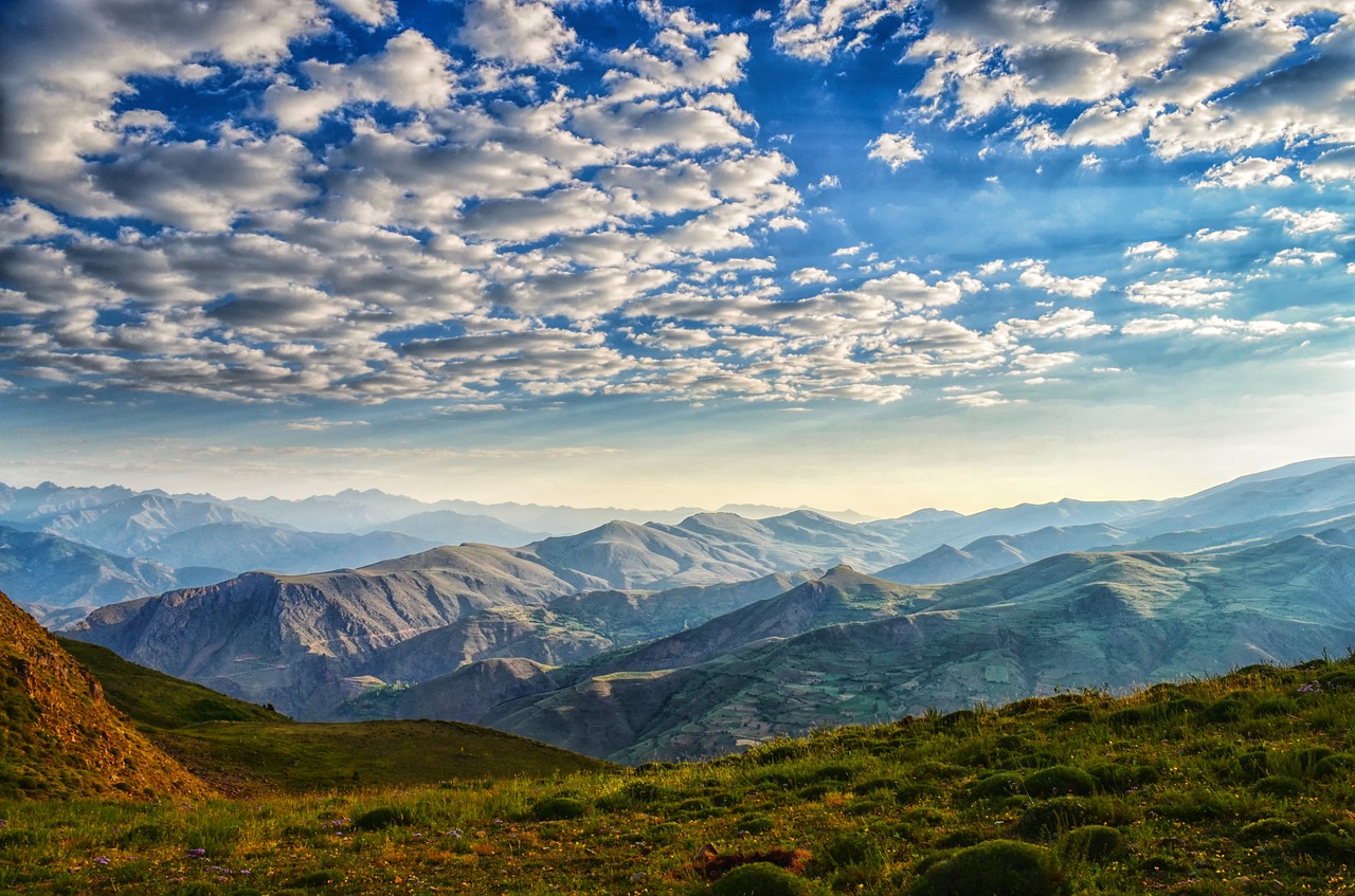 A vast mountainous landscape under a sky filled with scattered clouds. The foreground features green hills and vegetation.