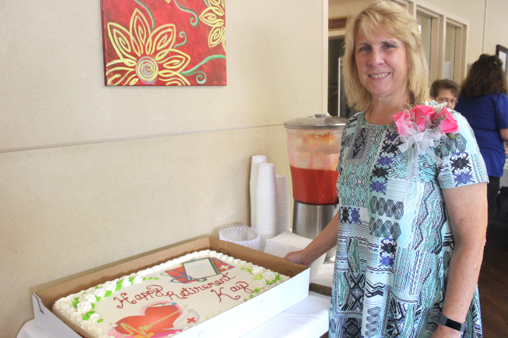 A woman with a corsage stands beside a decorated cake with "Happy Retirement" written on it. As the director of nursing, she beams with pride. There is a beverage dispenser and a painting in the background.