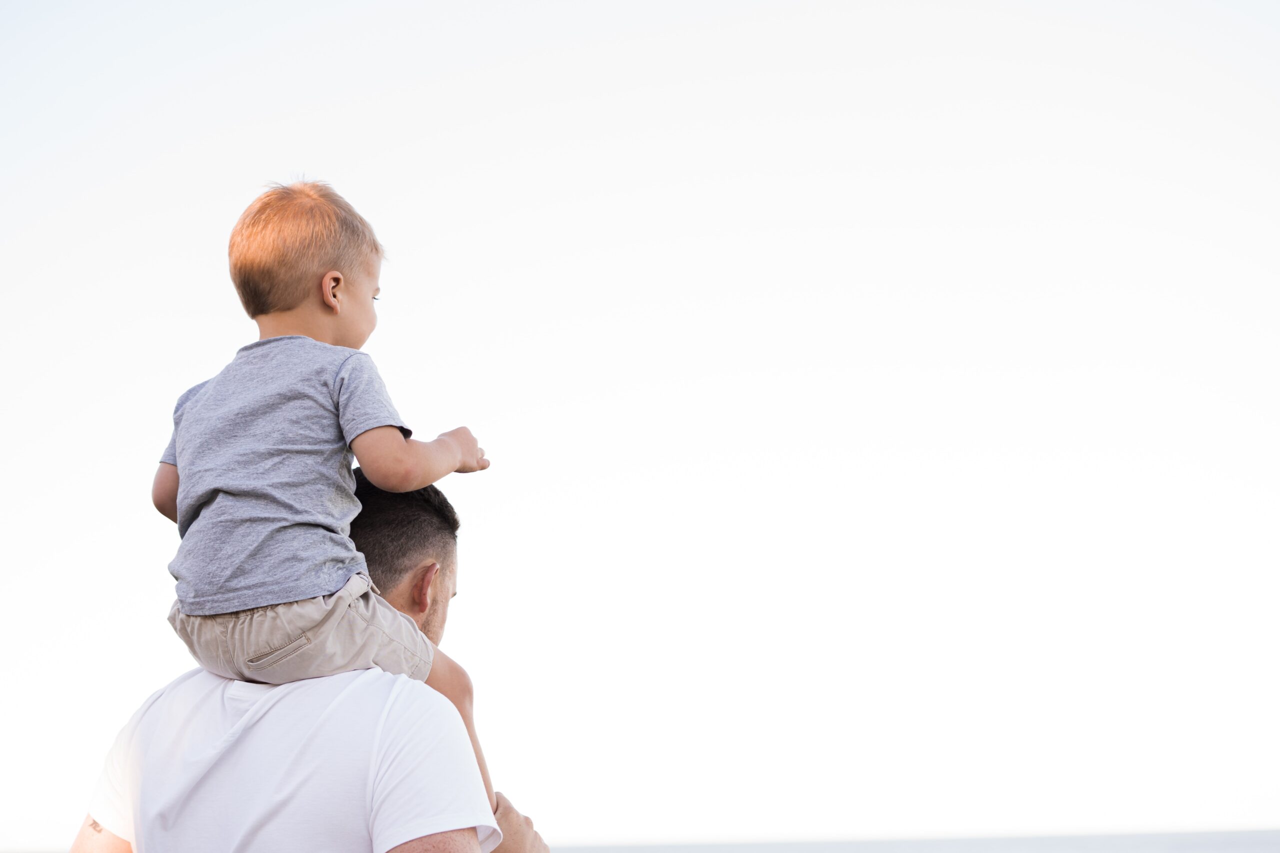 An adult carries a young child on their shoulders while both face towards the background under a clear sky.