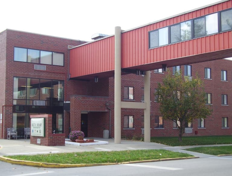 A three-story brick building in Martinsville, Indiana, with a covered skywalk connecting the top floor to another structure, featuring a sign, small garden area, and entrance doors on the ground floor. This is an example of affordable housing designed to blend functionality with charm.