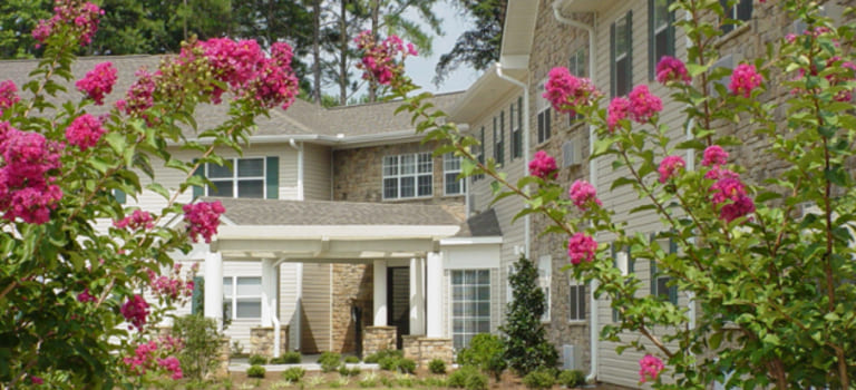 An exterior view of affordable housing in Kennesaw, Georgia, with flowering pink shrubs in the foreground and a pathway leading to the entrance.