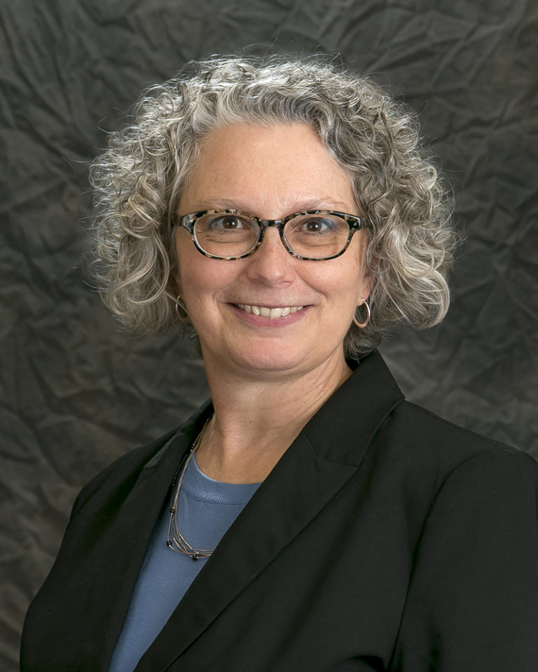 A person with curly gray hair, wearing glasses, a black blazer, and a blue shirt, is smiling with an air of leadership in front of a dark, textured background.