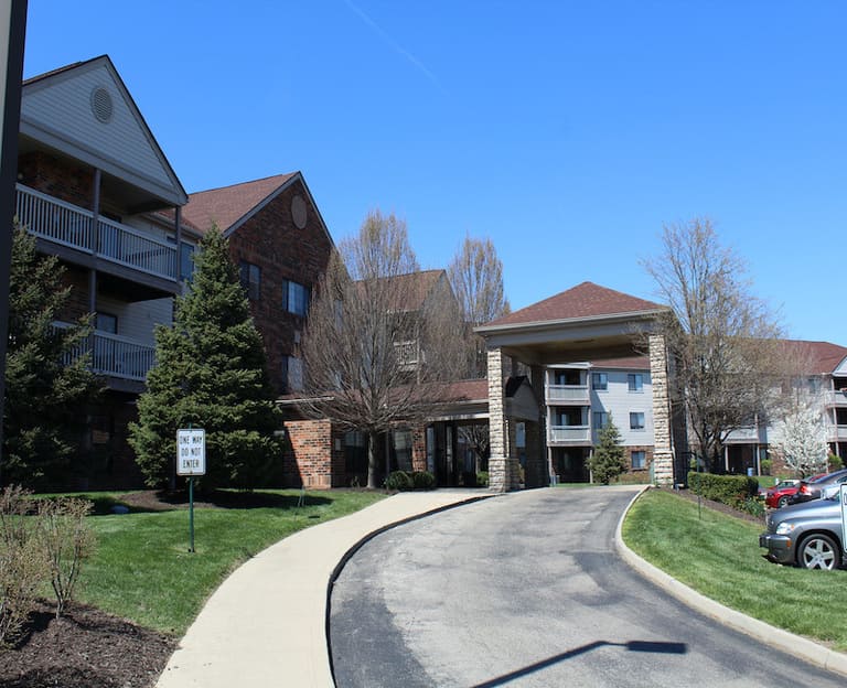 A curved driveway leads to the entrance of a multi-story residential building with a porch. Manicured lawns and trees surround the area, offering serene surroundings for affordable housing in Dayton, Ohio.