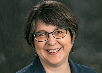 Woman with short brown hair and glasses smiling against a dark background, wearing a dark shirt and earrings.