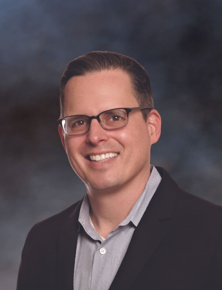 A person with short dark hair and glasses, wearing a striped shirt with a dark blazer, smiles confidently against a dark, blurred background, exuding an air of leadership.