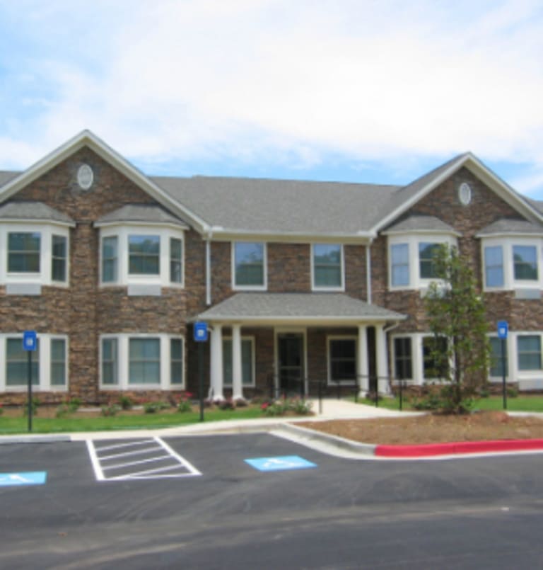 A two-story brick building with multiple windows, a covered entrance, and a concrete pathway. The front features a small garden, with parking spaces in the foreground, including accessible parking. This charming structure adds to the landscape of affordable housing in Jackson, Georgia.