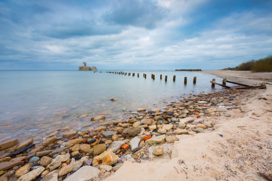 A sandy beach with scattered rocks and remnants of wooden posts leading into calm sea water. An old, abandoned structure is visible in the distance under a cloudy sky.