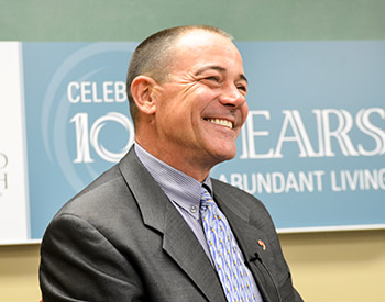 Man in a suit smiling in front of a backdrop with text reading "Celebrating 100 Years" and "Abundant Living.