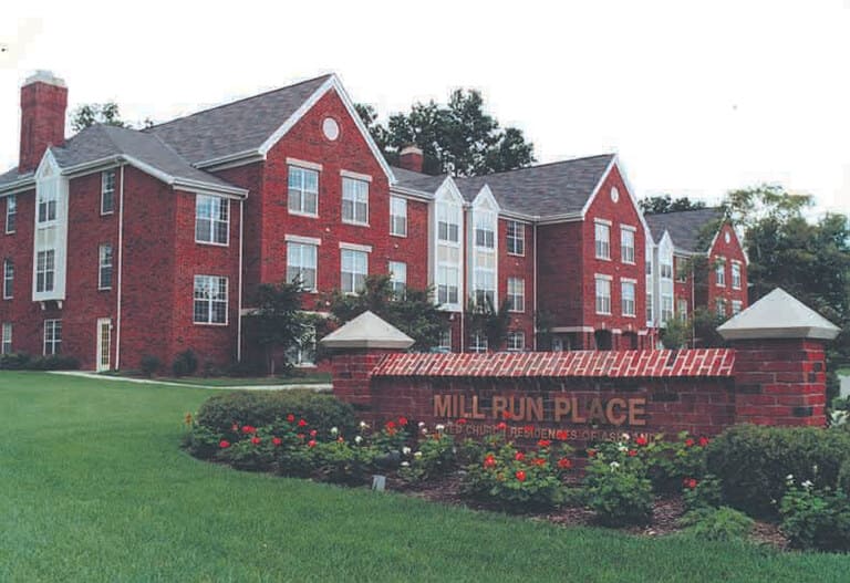 Image of an apartment complex with brick buildings labeled "Mill Run Place" on a brick sign in front, showcasing affordable housing in Ashland, Ohio, surrounded by a landscaped lawn and flowers.