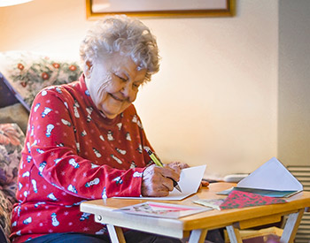 An elderly woman with curly white hair sits at a small table, writing on a card. She is wearing a red sweater with a snowman pattern and smiling.