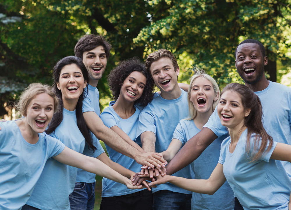 A diverse group of young people in blue shirts stand outdoors in a circle, smiling and stacking their hands together in the center, embodying uch company values. Trees and greenery are visible in the background.