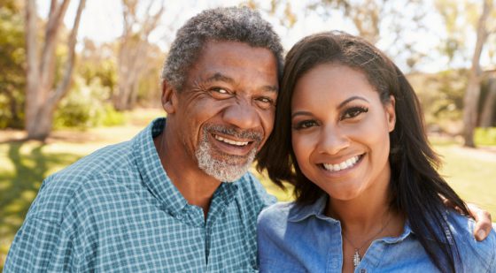 An older man and a younger woman, both smiling, stand close together outdoors on a sunny day with trees in the background, representing the caring community spirit fostered by United Church Homes.