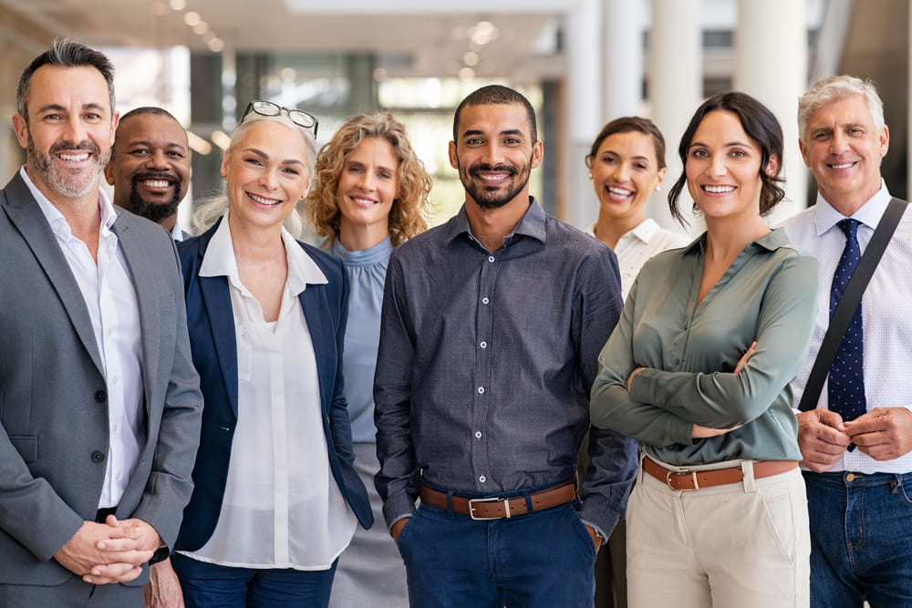 A diverse group of eight people, dressed in business casual attire, stands together in a bright, modern office space, smiling at the camera. This image embodies the welcoming and inclusive environment that answers the question: why work at United Church Homes?