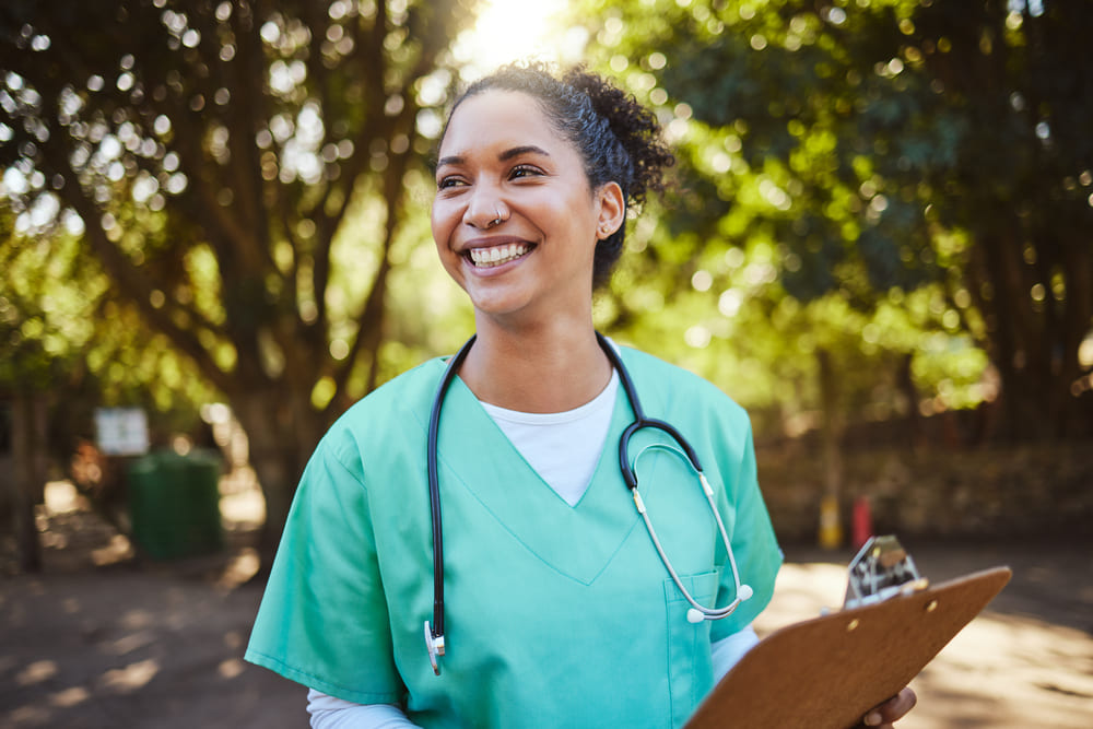 A smiling healthcare worker wearing scrubs and a stethoscope holds a clipboard outdoors with sunlight filtering through trees in the background, showcasing the rewarding nature of nursing jobs.