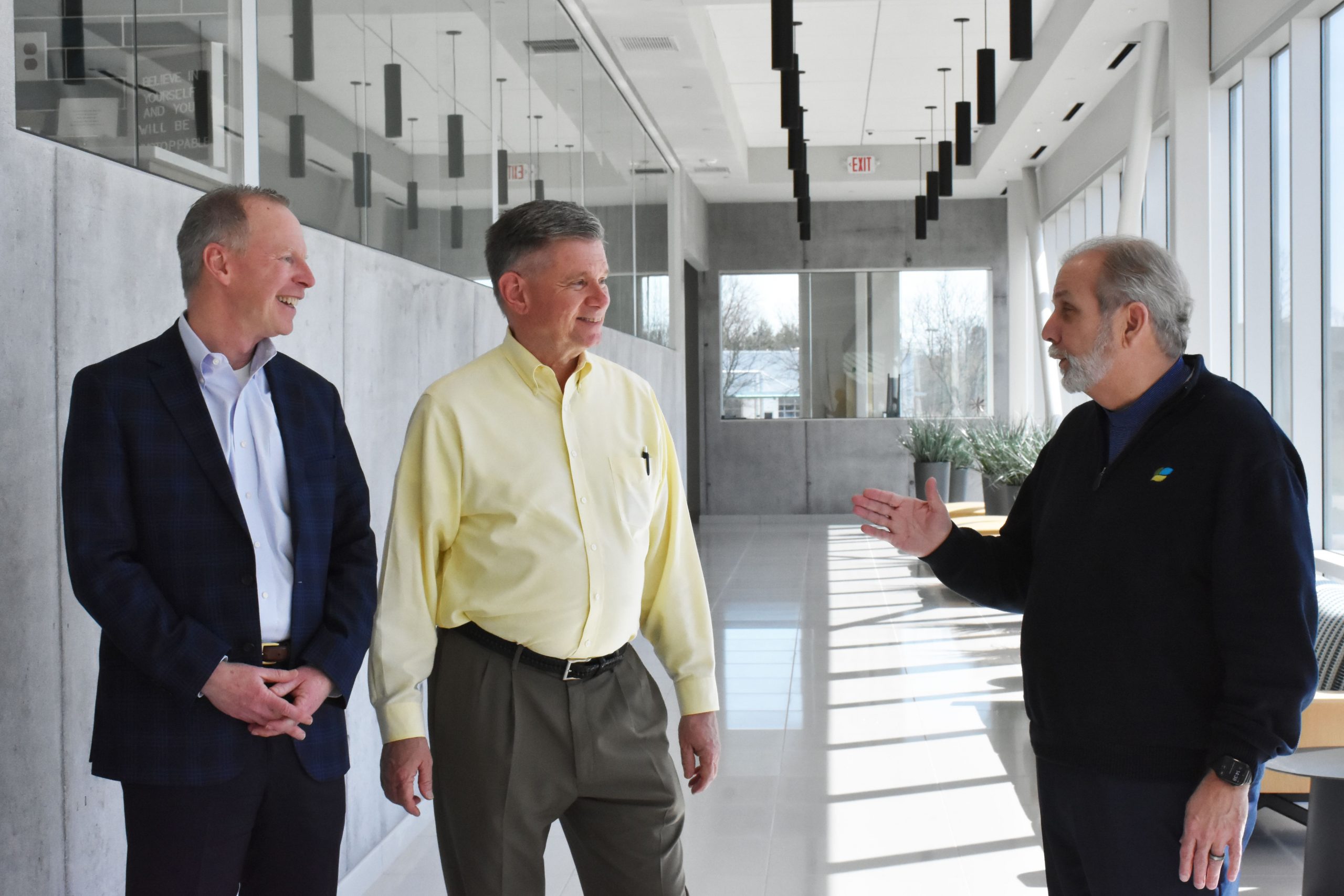 Three men are standing and conversing in a bright, modern hallway with large windows. One person gestures with his hand while the others listen intently, embodying the spirit of transformational change as the Radiant Alliance unites to reshape the healthcare landscape.