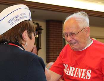 An elderly man in a red "Marines" shirt is emotional while interacting with a woman wearing a hat labeled "Department of 1st Vice President.