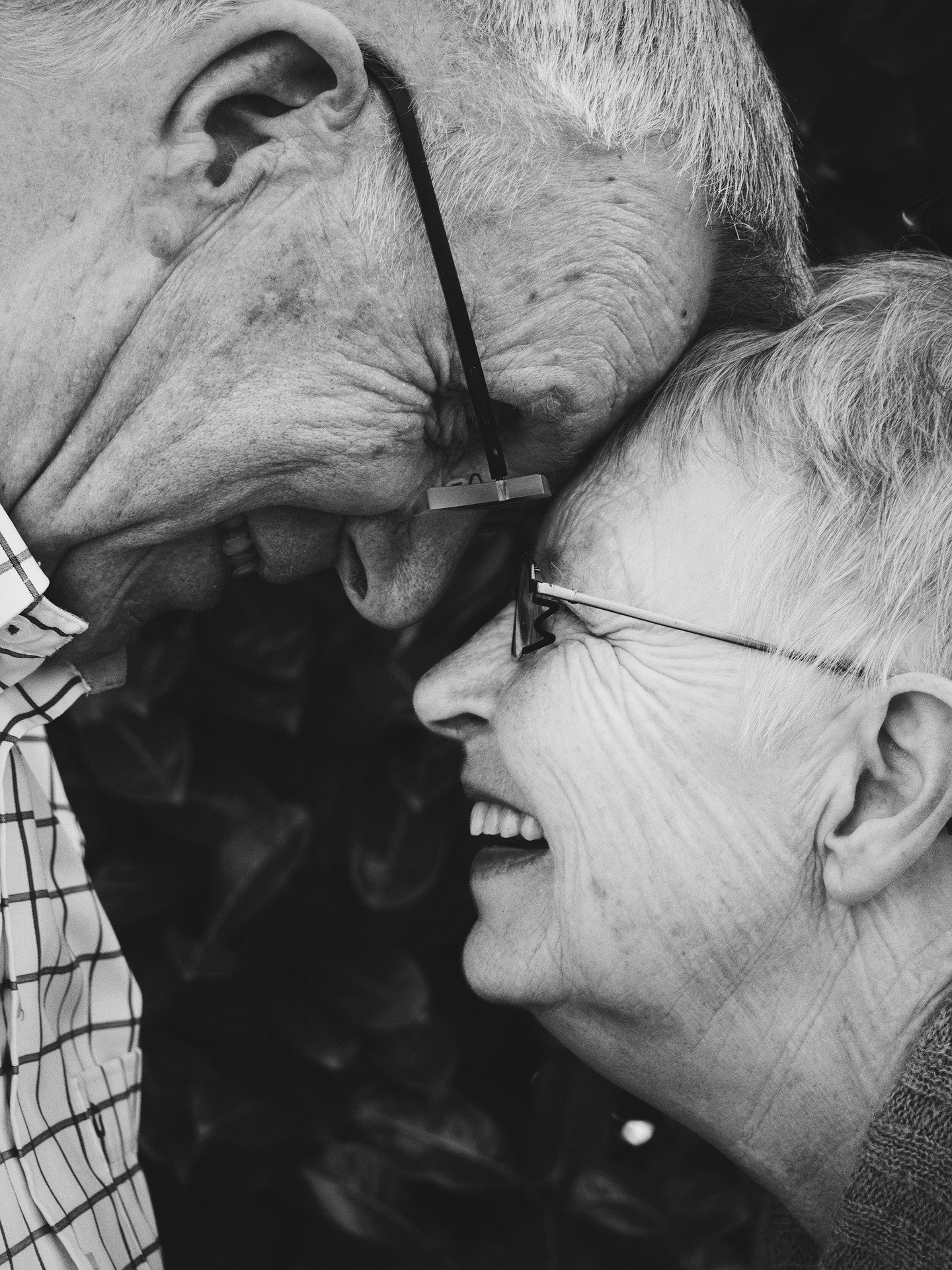 Two elderly people, one wearing a checkered shirt and the other in glasses, smile while touching foreheads, surrounded by dark foliage background.