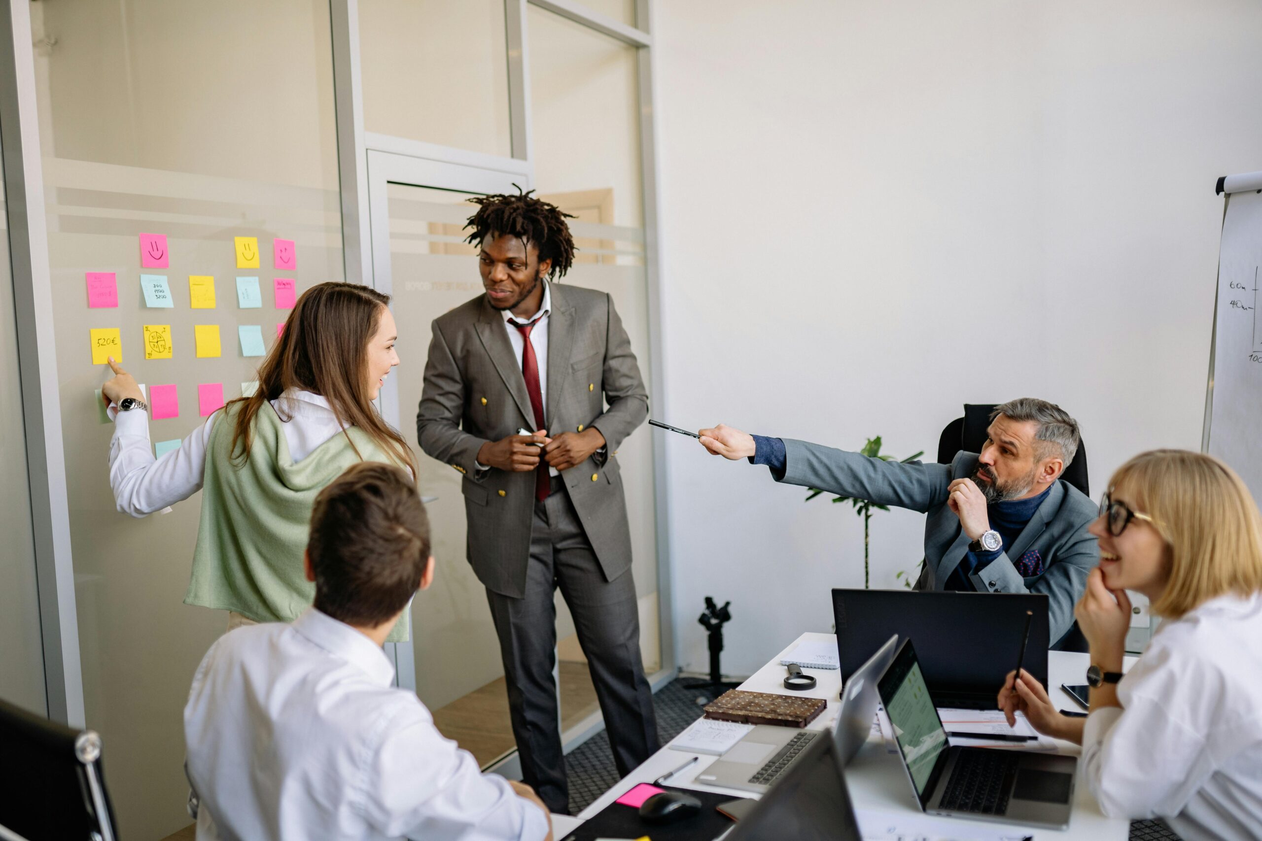 A group of colleagues engage in a discussion in a meeting room, with sticky notes on a glass wall, laptops on the table, and a flip chart in the background, strategizing on senior living community management.