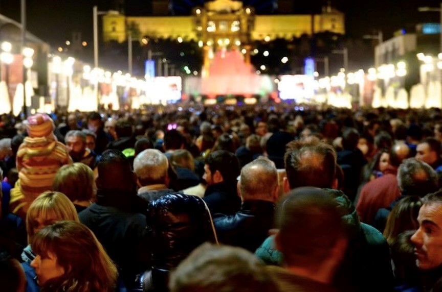 A large crowd gathers at night in front of an illuminated building, with many people facing the stage or focal point ahead. The scene is vibrant and busy with bright lights in the background.