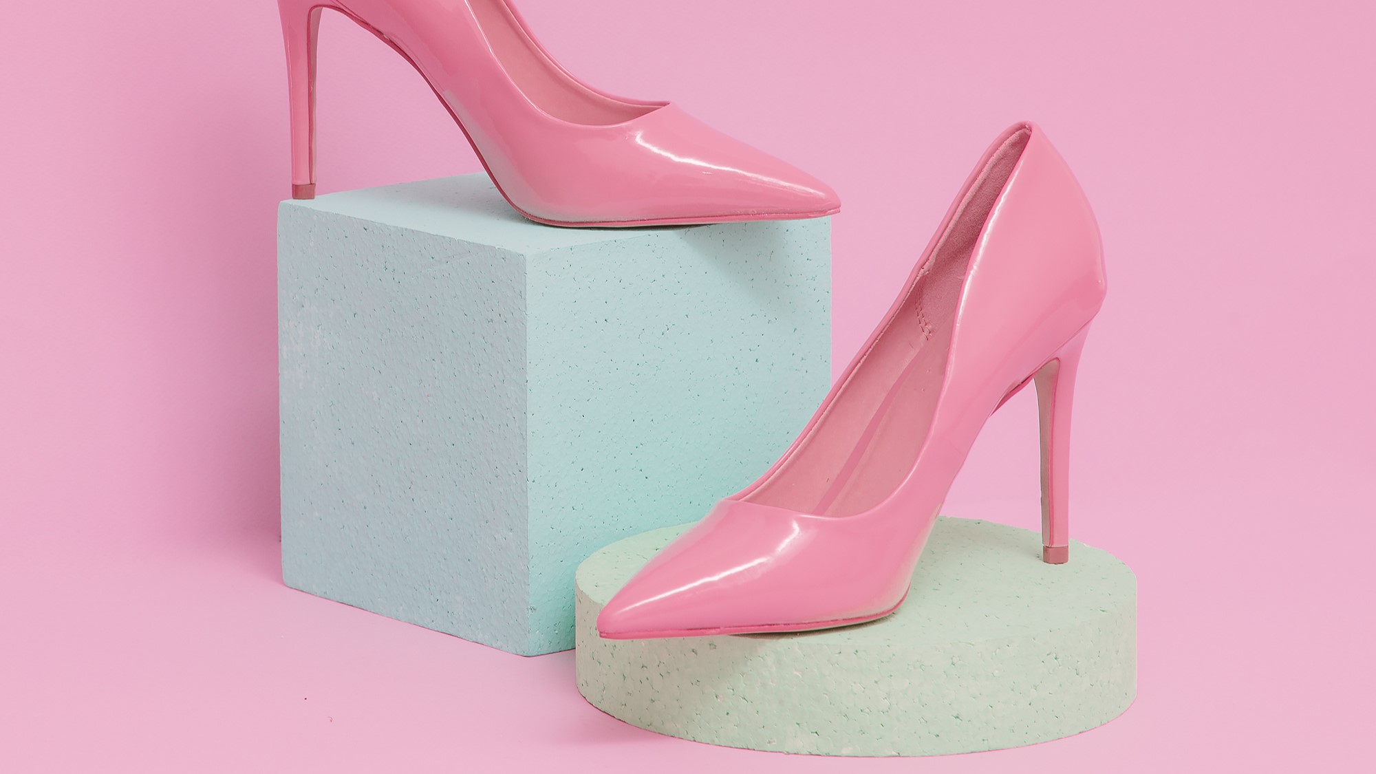 Two pink high-heeled shoes are displayed on blue foam-like pedestals against a pink background.