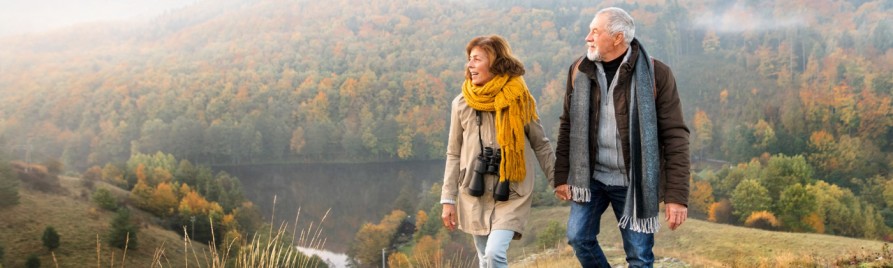 An older couple, dressed warmly, walks hand in hand on a hill overlooking a forested valley with autumn foliage. The woman has binoculars around her neck and they seem to be enjoying the scenery.