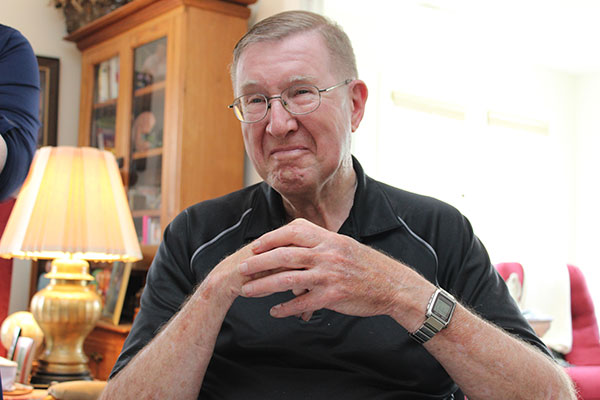 An elderly man wearing glasses and a black polo shirt is seated indoors, clasping his hands. Behind him are a lamp and a wooden cabinet with glass doors.