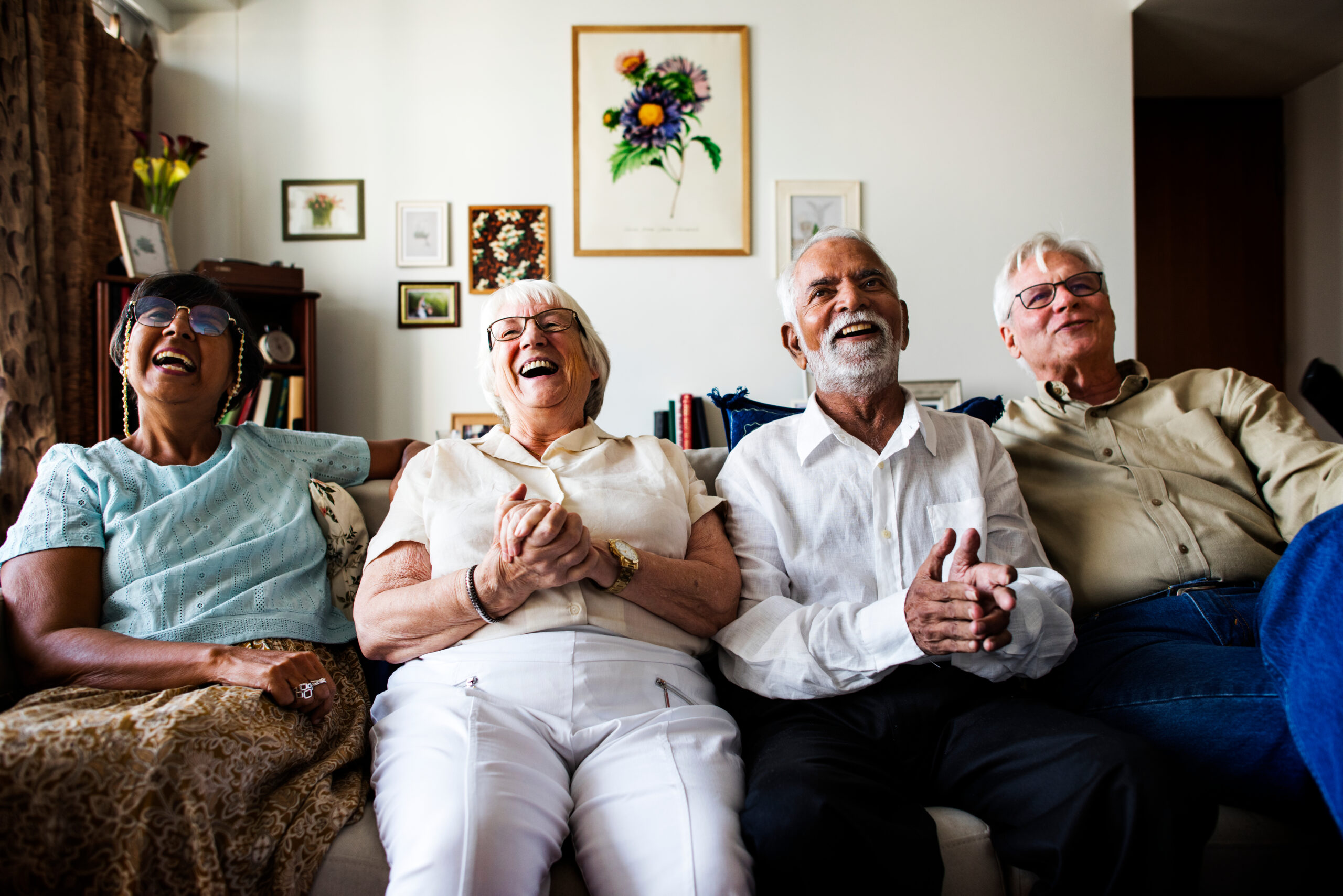 A group of four elderly people sits on a couch, laughing and smiling. They appear to be enjoying a moment together in a cozy living room decorated with framed pictures and flowers.