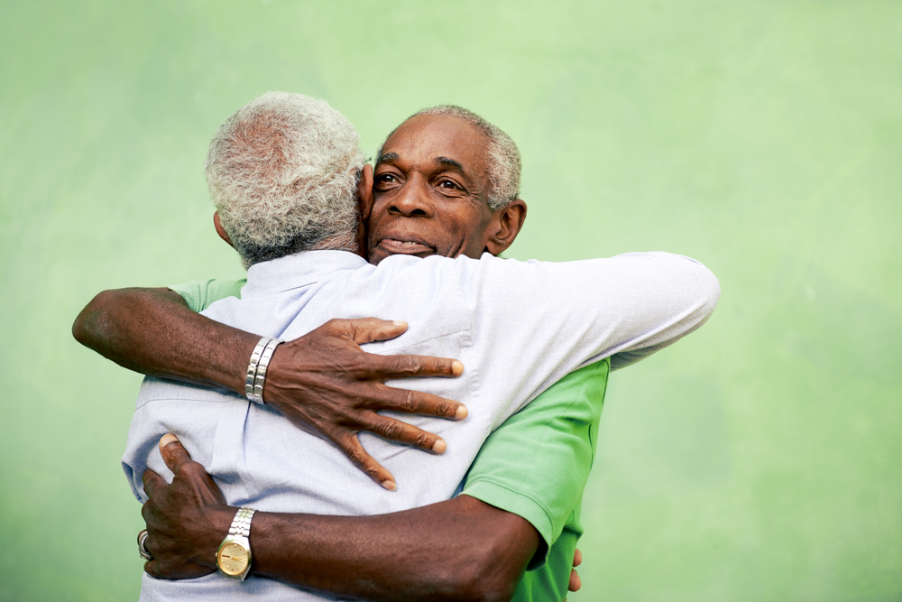 Two elderly men with gray hair are seen embracing warmly against a plain, green background. One man is wearing a green shirt and the other man is wearing a light blue shirt.