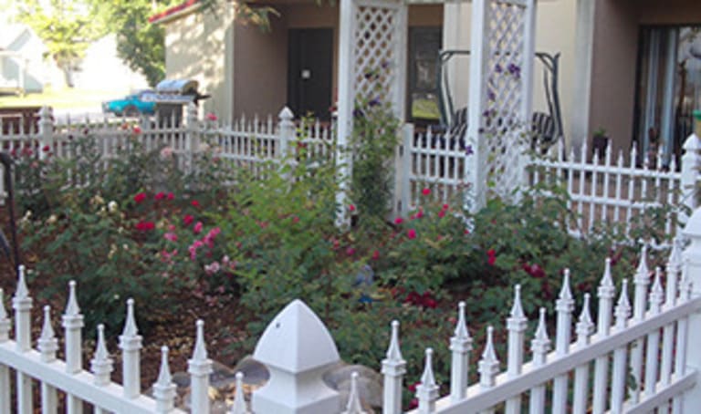 A small garden with red roses is enclosed by a white picket fence, adding charm to the affordable housing in Lewistown, Illinois. Two white trellises are in the garden, and a porch swing is visible in the background.