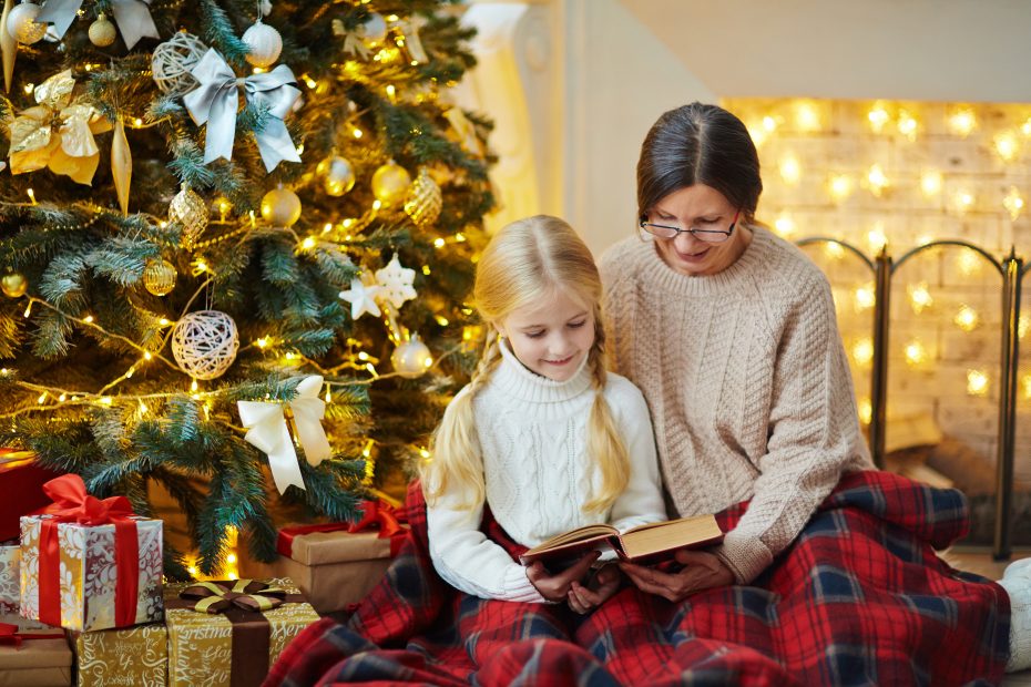 A woman and a child sit together, reading a book in front of a decorated Christmas tree with gifts underneath. They are wrapped in a red plaid blanket.