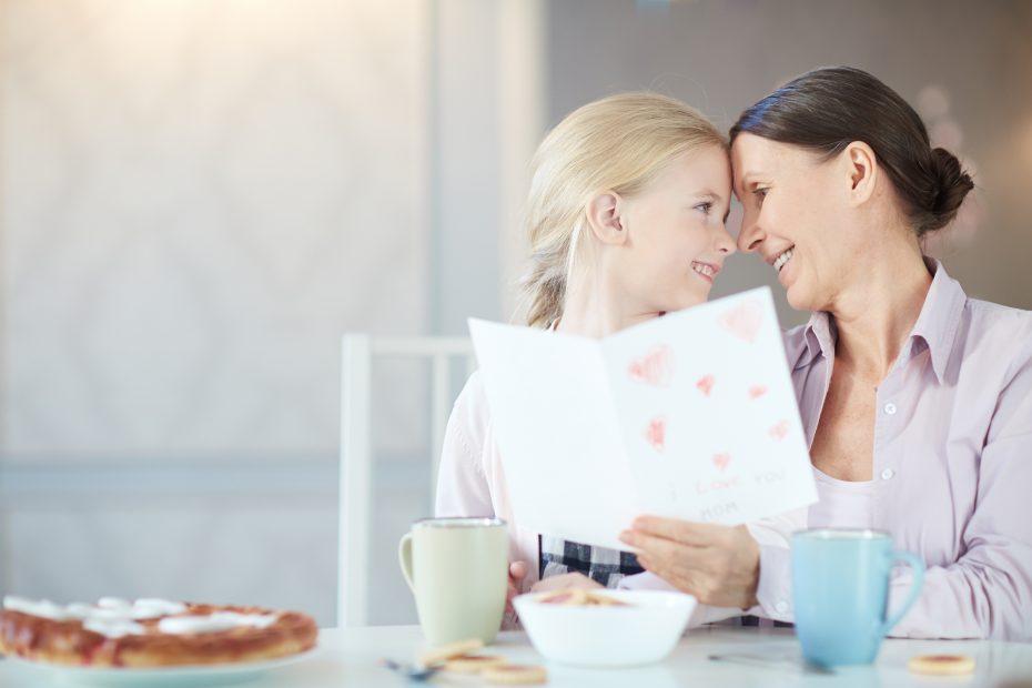 A woman and a child sitting at a table, sharing a close moment while holding a card with heart drawings. A pizza and mugs are on the table.