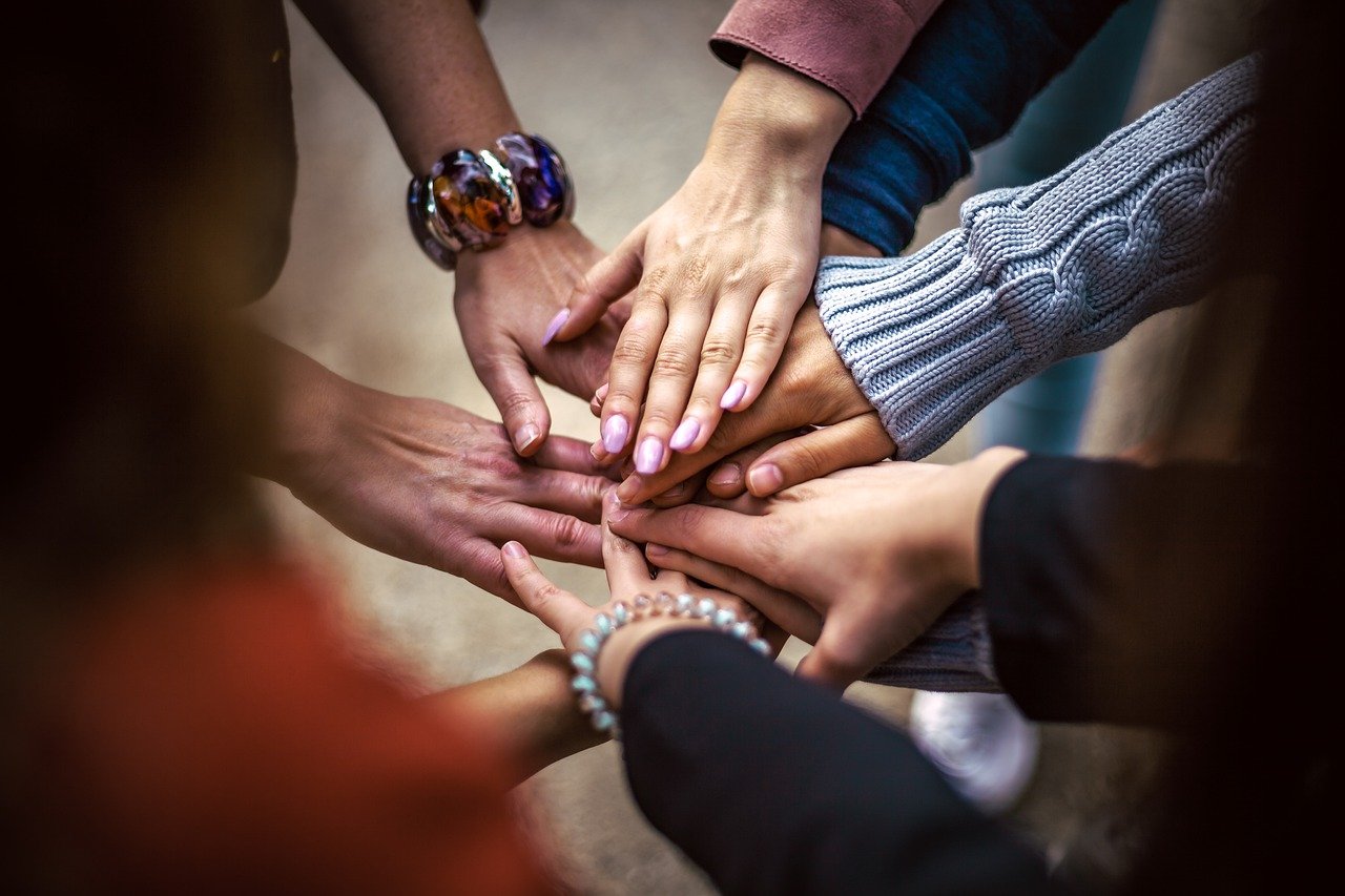 A group of people put their hands together in a stack, showing unity and teamwork. The photo focuses on the hands, with various skin tones and different kinds of clothing visible.