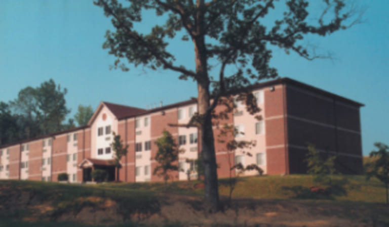 A three-story red brick building with white accents, surrounded by greenery and trees under a clear blue sky, offers affordable housing in Rome, Georgia.