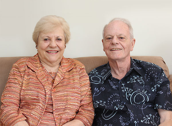 An older couple sitting on a couch, smiling at the camera. The woman wears an orange patterned jacket, while the man wears a black shirt with white designs.