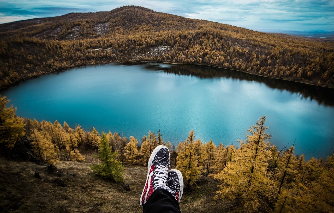 View of a person’s feet hanging over a cliff, with a blue lake surrounded by autumn-colored trees and hills in the background.