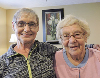 Two older women with glasses smile at the camera. The woman on the left wears a grey zip-up jacket, and the woman on the right wears a pink jacket with a blue collar. A floral painting hangs in the background.