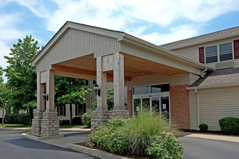 A single-story affordable housing unit in Dayton, Ohio, features a covered entrance supported by wooden pillars, surrounded by landscaped shrubs and trees, with a paved walkway leading to the glass entrance door.